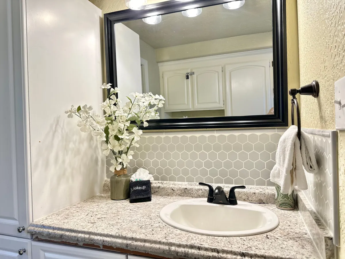 A bright and calming bathroom with a granite countertop, sink, mirror, neatly folded towels, and a fresh flower arrangement. The space exudes cleanliness and comfort, perfect for refreshing morning or evening routines.