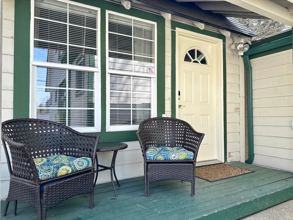 A cozy front porch scene with woven chairs and decorative cushions, positioned beside the front door. Sunlight fills the space, inviting residents or guests to relax outdoors, socialize, or enjoy a quiet moment before heading out.