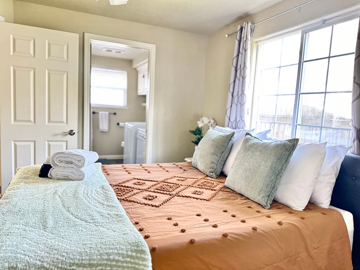 A serene bedroom filled with natural light, featuring a queen bed dressed in a decorative bedspread and pillows. The window allows sunlight to stream in, creating a cozy and restful atmosphere perfect for relaxation or quiet activities like reading or enjoying a morning coffee.