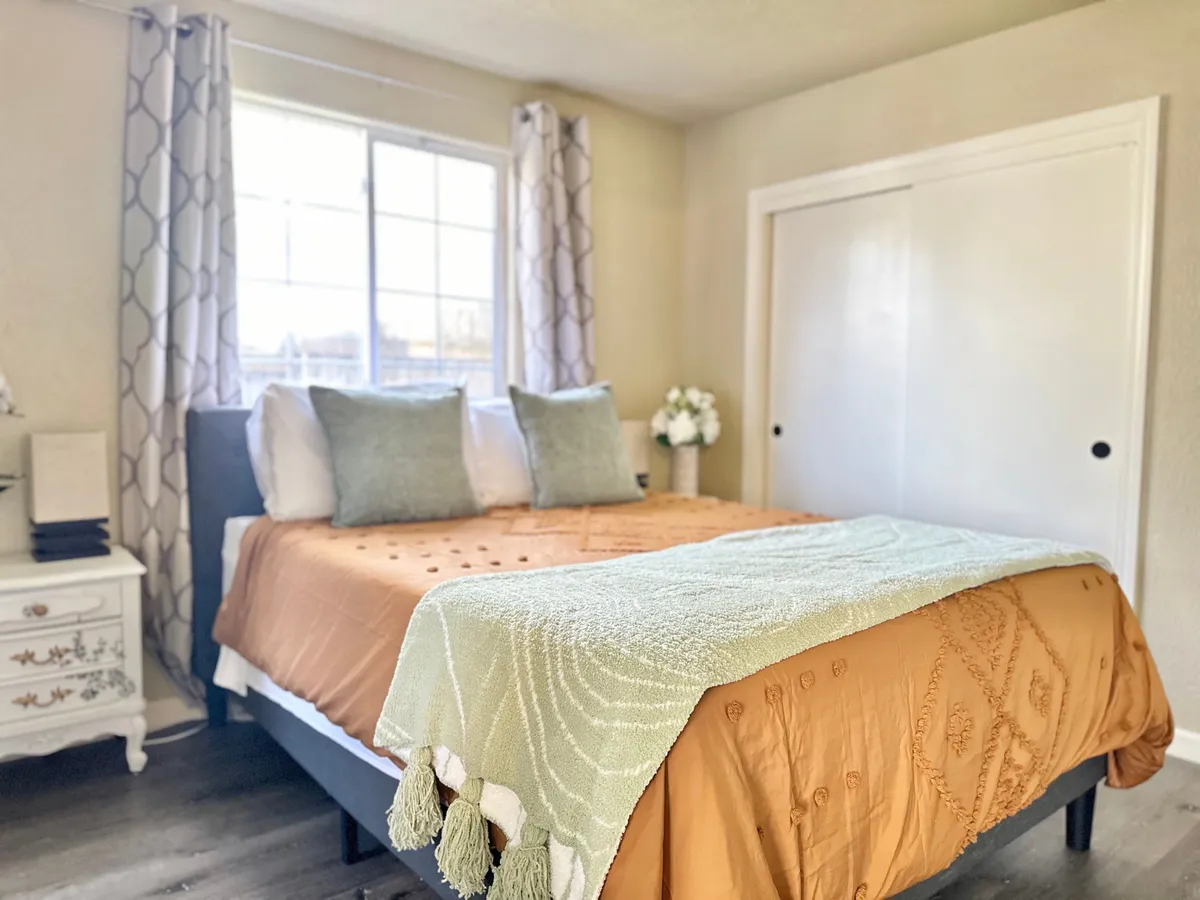 A cozy queen-sized bed dressed in decorative bedding, placed near a window that streams in sunshine. A nightstand with tasteful decor sits beside the bed, creating an inviting and restful atmosphere.