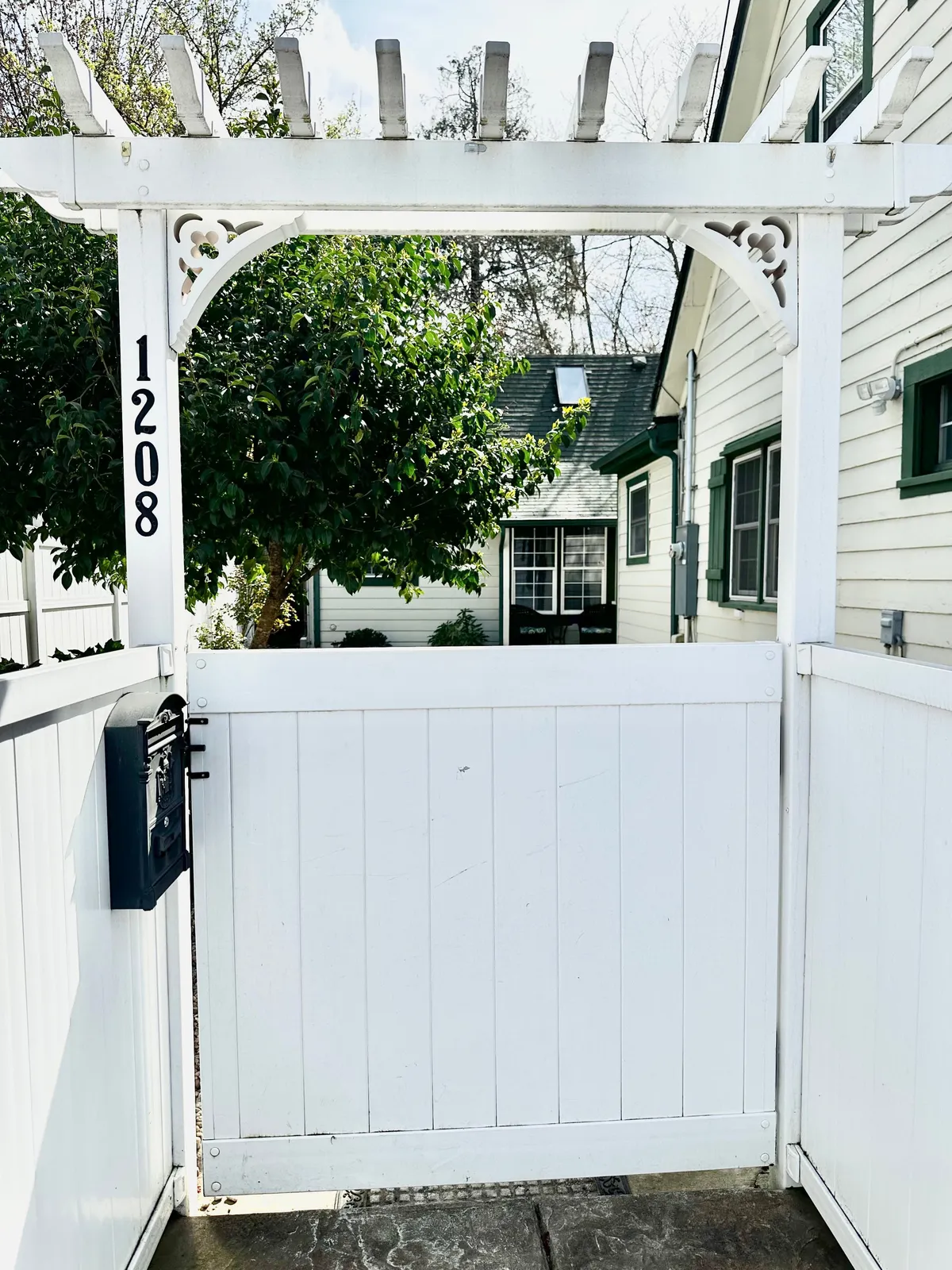 A serene exterior scene featuring a white gate with a pergola, lush green foliage, and the house in the background. The composition suggests a peaceful starting point for adventures into the city, while also conveying a sense of homecoming and community.