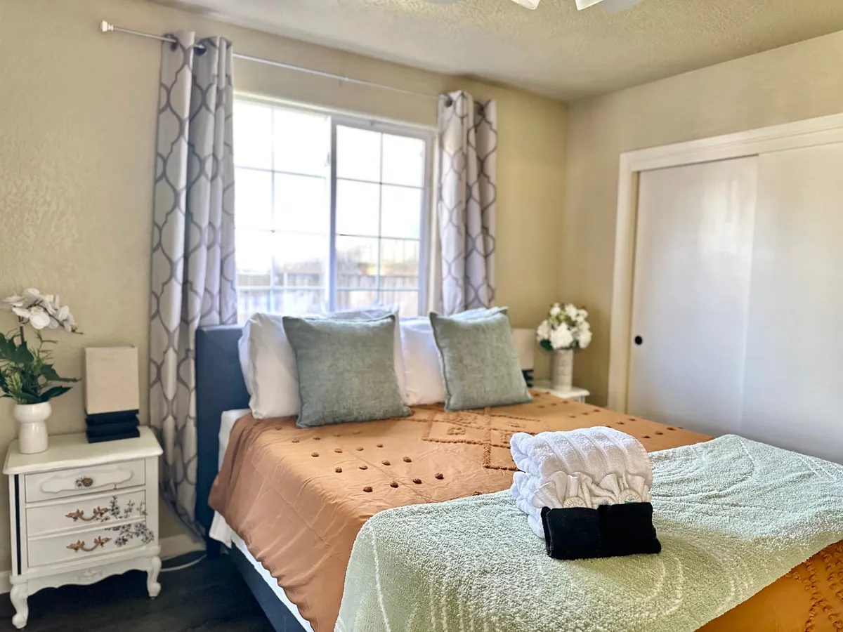 A welcoming bedroom scene showcasing a bed with decorative pillows, a nightstand adorned with fresh flowers, and a window with soft curtains. The overall mood is inviting and tranquil, offering a restful retreat for guests.