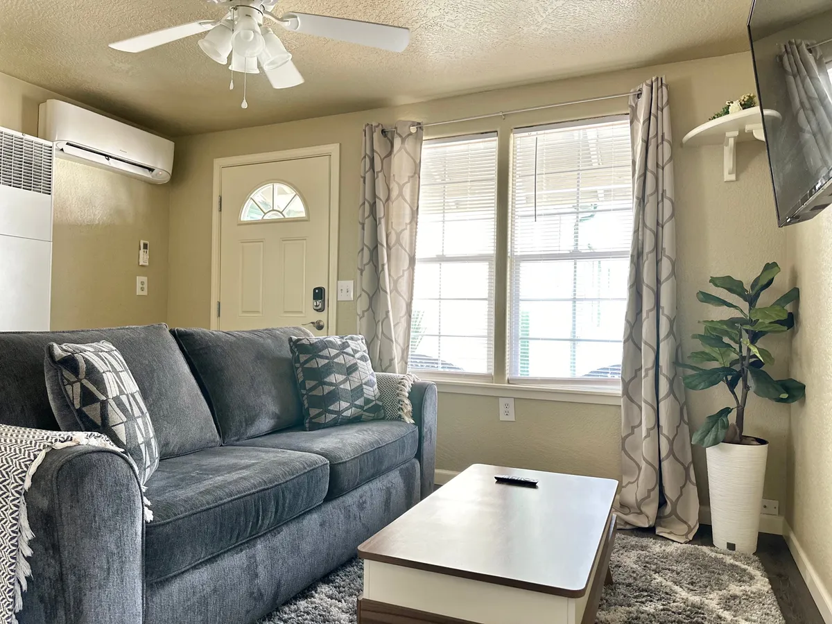 This image showcases a bright, inviting living area with a plush sofa, coffee table, large windows letting in natural light, and a touch of greenery from a nearby plant. The setup exudes comfort and relaxation—perfect for lounging, enjoying coffee, or spending time with friends and family.