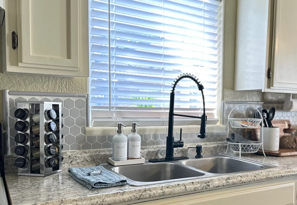 A bright kitchen area with a modern faucet, spice rack, kitchen sink, and soap dispensers, illuminated by natural light through a window with blinds. The scene is welcoming and functional, showcasing a clean space for meal preparation or winding down.