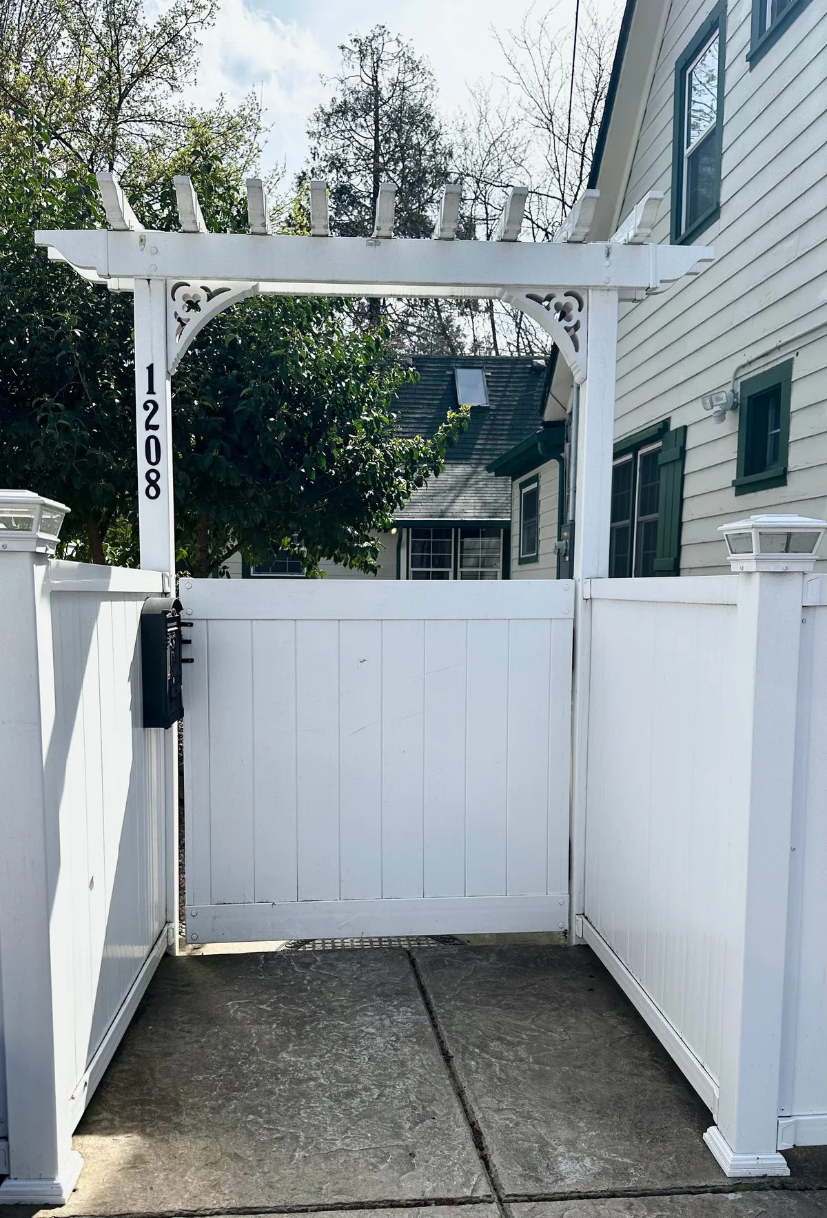 A welcoming white wooden gate set beneath a charming pergola marks the entrance to the property, with the house facade and address number visible in the background. The setting is inviting, hinting at a lively flow between the private guest house and the vibrant surroundings.